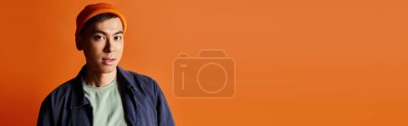 A stylish Asian man wearing a blue jacket and an orange hat poses against a vibrant orange background in a studio.