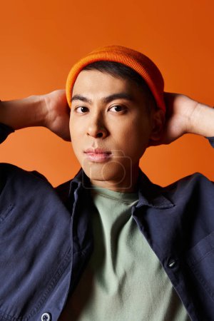 A handsome Asian man in a blue jacket and orange hat, exuding style and elegance against a vibrant orange background.