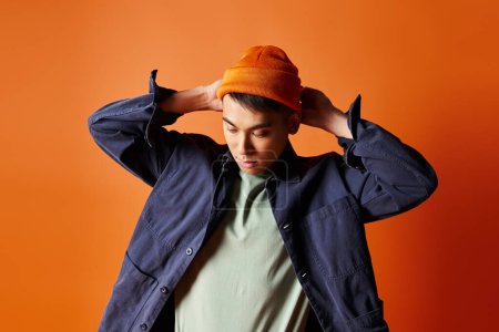 Ein gut aussehender asiatischer Mann in stilvoller Kleidung trägt selbstbewusst einen Hut auf dem Kopf vor orangefarbenem Hintergrund.