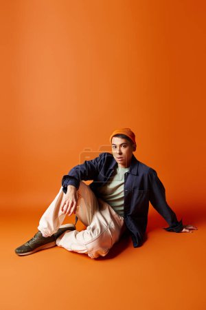 Foto de Un hombre asiático con estilo en un atuendo elegante sentado pacíficamente en el suelo sobre un fondo naranja. - Imagen libre de derechos
