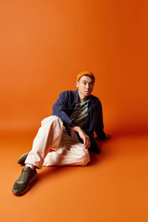 Schöner asiatischer Mann in stylischem Outfit sitzt auf orangefarbenem Hintergrund, tief in Gedanken oder Reflexion.
