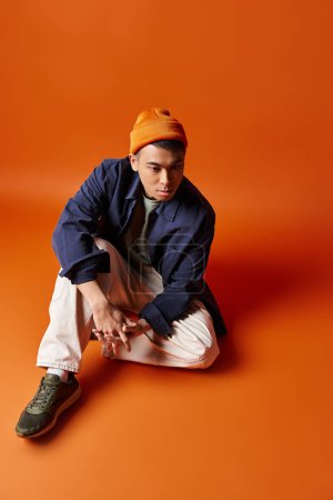 A fashionable young Asian man sits on the ground, wearing a trendy hat, exuding a contemplative vibe on an orange background.