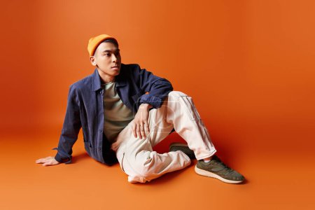 A handsome Asian man in stylish attire sits on the ground wearing a hat, against an orange background in a studio setting.