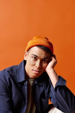 Handsome Asian man dressed in a blue shirt and an orange hat, standing confidently against a bright orange background in a studio.