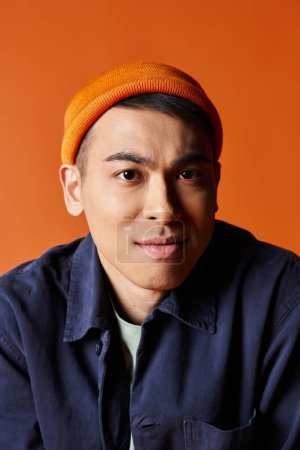 A handsome Asian man in a blue shirt and an orange hat stands confidently against an orange background in a studio.