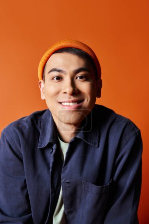 A handsome Asian man exudes style in a blue shirt and orange hat against a vibrant orange backdrop.