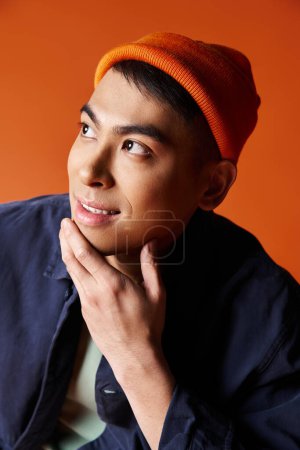 Un hombre asiático guapo lleva con confianza una chaqueta azul y un sombrero naranja sobre un fondo vibrante.
