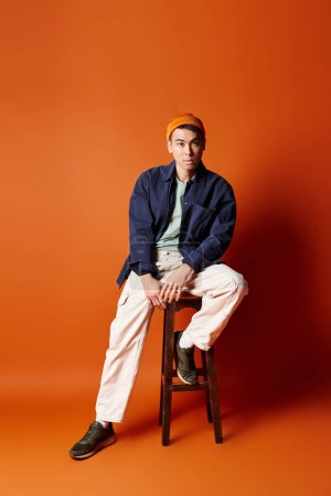 Ein gut aussehender asiatischer Mann in stylischer Kleidung sitzt selbstbewusst auf einem Holzhocker vor orangefarbenem Hintergrund..