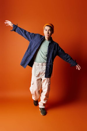 A handsome Asian man in a blue jacket and khaki pants poses against an orange background in a studio setting.