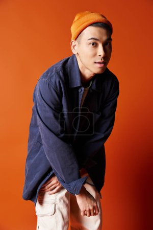 A handsome Asian man exudes style in a blue shirt and orange hat against a vibrant orange background. Poster 700749270