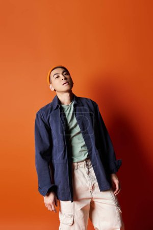 A handsome Asian man stands confidently in front of a striking orange wall in a stylish outfit.