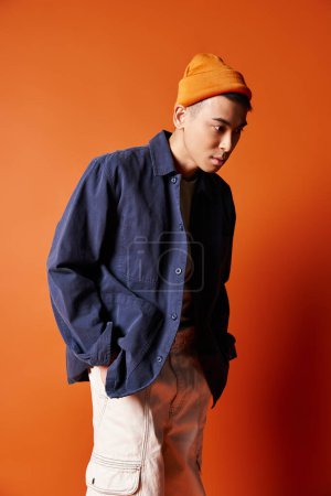 Un joven asiático elegante con una camisa azul y un sombrero naranja posa con confianza sobre un fondo naranja vibrante.