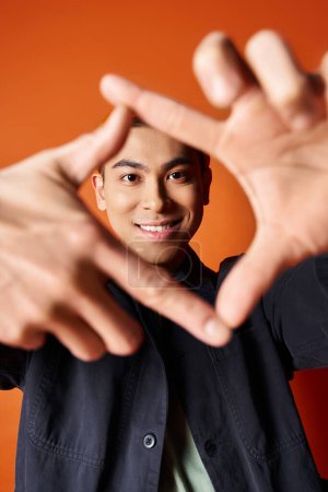 Handsome Asian man in stylish attire shapes a heart with his hands against an orange studio backdrop.