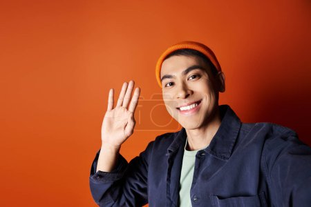 Handsome Asian man in blue jacket and orange hat confidently waving hand against a bold orange background.