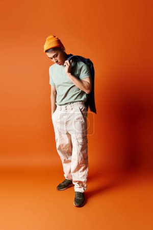 A handsome Asian man confidently poses in a green shirt and white pants against an orange background in a studio setting.