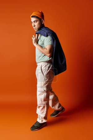 A handsome Asian man in stylish attire carries a backpack on his back against an orange background in a studio setting.