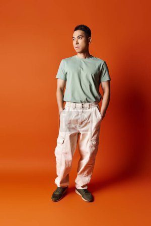 A handsome Asian man stands confidently in stylish attire against a bright orange background in a studio setting.