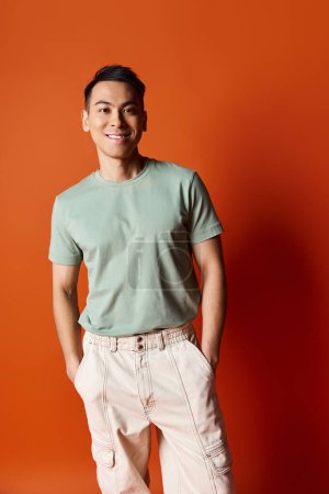 A fashionable Asian man in stylish attire standing confidently in front of a vibrant orange wall in a studio setting.