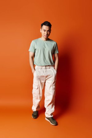 Un hombre asiático guapo se para con confianza frente a una vibrante pared naranja, emanando un sentido del estilo y la individualidad.