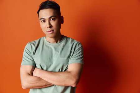 A handsome Asian man stands confidently with crossed arms against a striking orange wall in a stylish studio setting.
