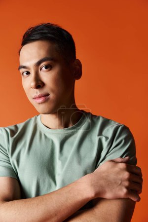 Handsome Asian man with crossed arms poses confidently in stylish attire against orange studio backdrop.