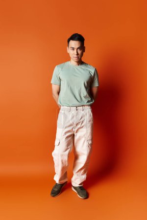 Handsome Asian man in stylish attire standing confidently in front of a vibrant orange backdrop in a studio.