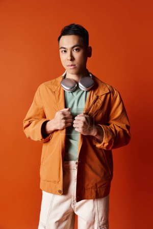 A stylish Asian man in an orange jacket and white pants striking a confident pose against an orange backdrop in a studio.