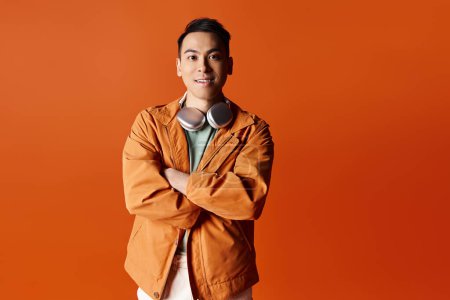 Handsome Asian man in stylish attire stands with arms crossed in front of vibrant orange background in a studio setting.