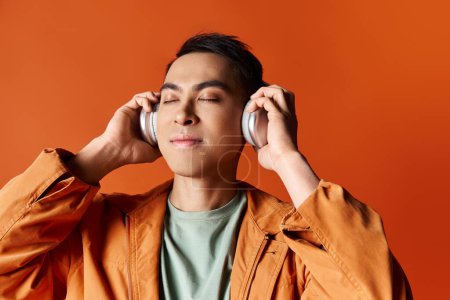 A handsome Asian man in stylish attire holding headphones to his ears on an orange background.