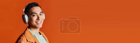 Photo for A handsome Asian man in stylish attire wearing headphones and a jacket, captured against an orange studio background. - Royalty Free Image