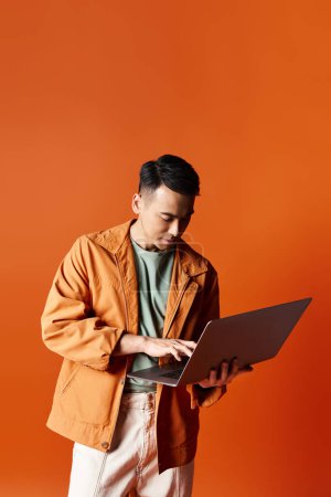Téléchargez les photos : Un homme asiatique élégant dans une veste orange absorbé par le travail sur un ordinateur portable dans un cadre de studio. - en image libre de droit