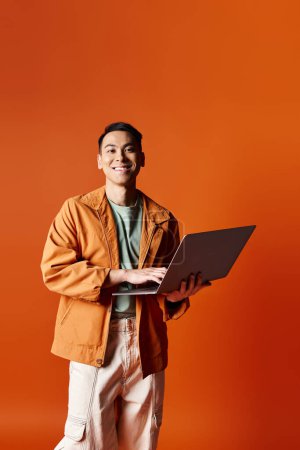 Foto de Un hombre asiático guapo, vestido con un atuendo elegante, sosteniendo un ordenador portátil mientras está de pie sobre un fondo naranja en un estudio. - Imagen libre de derechos