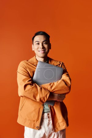 Un hombre asiático elegante con los brazos cruzados, sosteniendo con confianza un ordenador portátil contra un telón de fondo naranja.