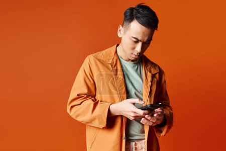 Téléchargez les photos : Un bel homme asiatique dans une veste orange élégant absorbé dans son téléphone portable. - en image libre de droit
