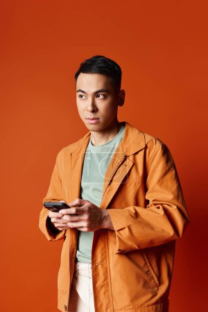 A handsome Asian man in a stylish orange jacket holding a cell phone against an orange background in a studio setting.