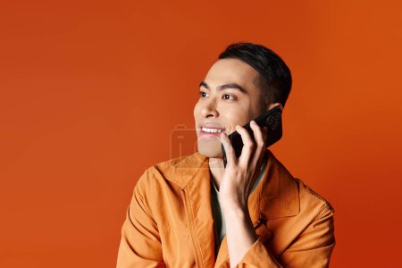 Handsome Asian man in stylish orange shirt engaged in a phone conversation against vibrant background.