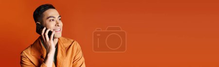 Ein stilvoller asiatischer Mann unterhält sich auf einem Handy vor einem grellen orangefarbenen Hintergrund.