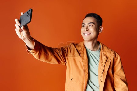 glücklicher asiatischer Mann in stylischer Kleidung, der ein Selfie mit seinem Handy vor orangefarbenem Studiohintergrund macht.