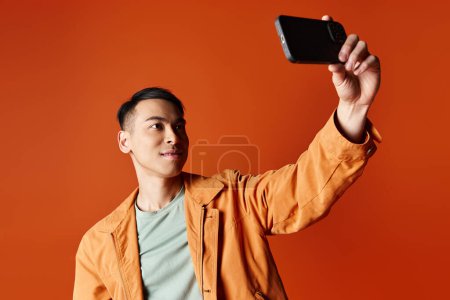 Schöner asiatischer Mann in stylischer Kleidung macht ein Foto mit seinem Handy vor orangefarbenem Studiohintergrund.