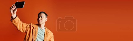 Ein gut aussehender asiatischer Mann in stylischer Kleidung hält vor orangefarbenem Hintergrund ein Handy in die Luft.