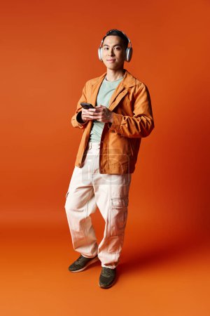 A stylish Asian man stands in front of a bold orange background, wearing headphones.