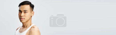 Téléchargez les photos : Un bel homme asiatique dans un débardeur blanc frappe une pose confiante dans un cadre de studio gris. - en image libre de droit