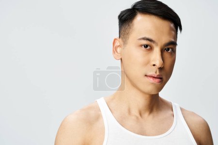 A young Asian man in a tank top strikes a pose in a grey studio setting, exuding confidence and tranquility.