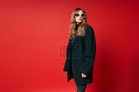 Téléchargez les photos : Belle femme à la mode en tenue élégante avec des lunettes de soleil posant sur fond rouge vibrant - en image libre de droit