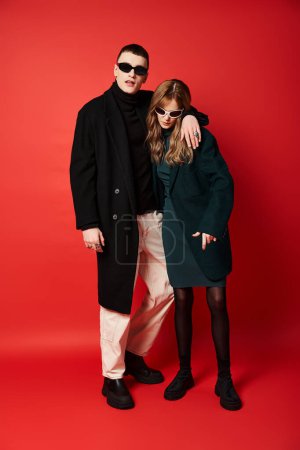 Photo for Fashionable beautiful couple in stylish coats with sunglasses posing together on red backdrop - Royalty Free Image