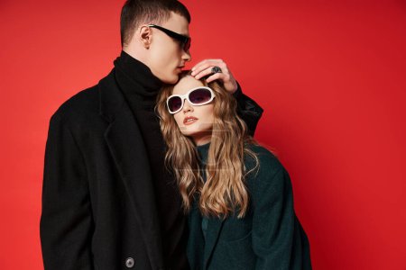 pareja joven de moda en abrigos elegantes con gafas de sol de moda posando juntos sobre el telón de fondo rojo