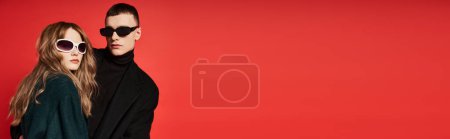 Photo for Loving young couple in stylish coats with sunglasses posing together on red backdrop, banner - Royalty Free Image