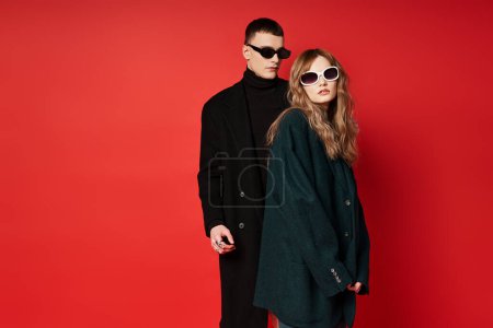 well dressed young couple in stylish coats with sunglasses posing together on red backdrop