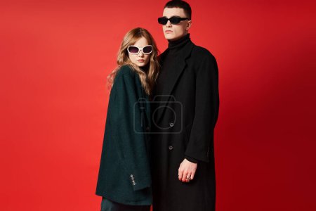 Téléchargez les photos : Élégant jeune couple en manteaux élégants avec des lunettes de soleil à la mode posant ensemble sur fond rouge - en image libre de droit