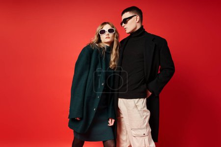 good looking young couple in stylish coats with sunglasses posing together on red backdrop
